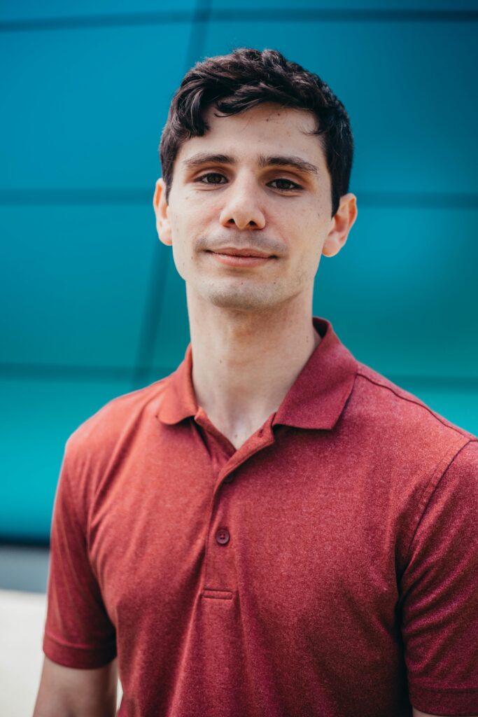 Portrait of a young man in a red polo shirt smiling outdoors against a teal backdrop.